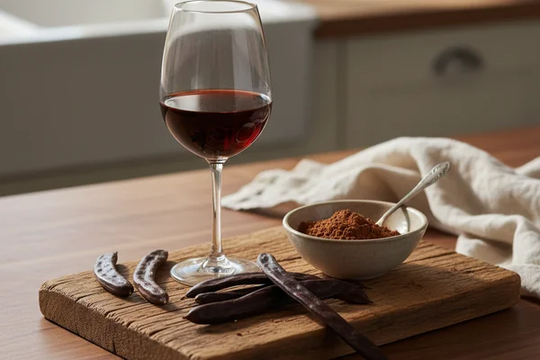 Carob pods and finished wine in a glass on a walnut surface bathed in warm natural light
