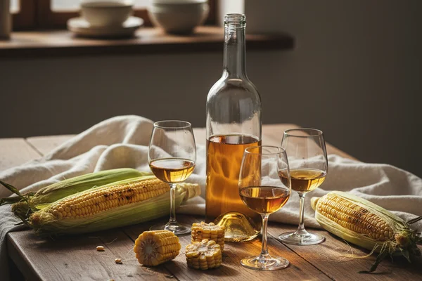 Rustic jar of golden corn wine beside dried corn cobs on a walnut surface in warm natural light
